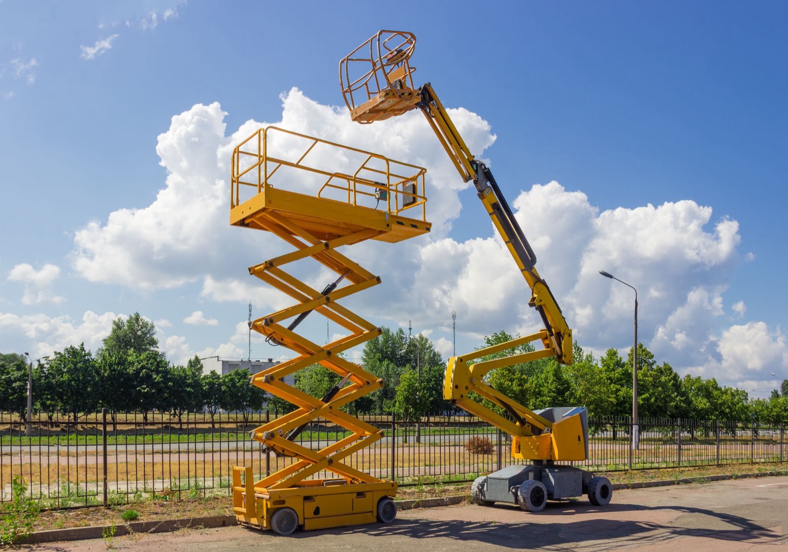A scissor lift and a boom lift with their platforms raised, parked beside a fence